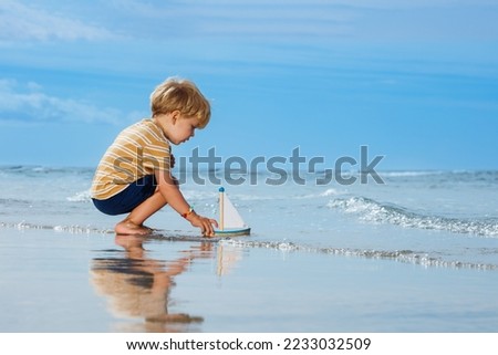 Happy handsome little blond boy put toy boat in the ocean waves at the beach during summer vacation