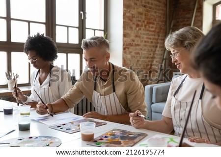 Happy handsome man in apron drawing picture with paints on paper, enjoying creative art master class workshop with smiling multiracial young and old people, sitting together at table in loft studio.