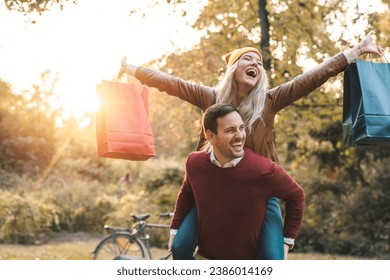 Happy handsome man carrying girlfriend on his back in a city park. Cheerful woman with arms outstretched holding shopping bags while piggybacking. - Powered by Shutterstock