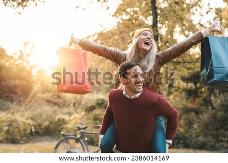 Happy handsome man carrying girlfriend on his back in a city park. Cheerful woman with arms outstretched holding shopping bags while piggybacking.