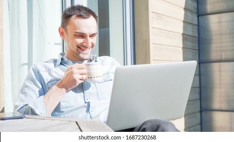 Happy Handsome Man Having A Facetime Video Call Talking To His Family. Smiling Casual Businessman Having A Video Conference Outside On A Sunny Day. Self Isolating At Home On Balcony Drinking Coffee. 