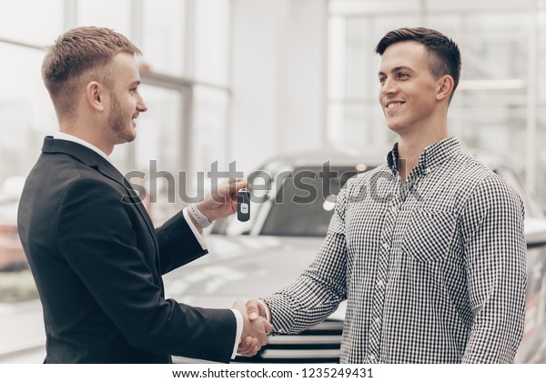 Happy handsome man receiving car keys to his new\
automobile from car salesman at the dealership. Professional auto\
seller shaking hands with his customer, handing him keys to a new\
vehicle. Buying car