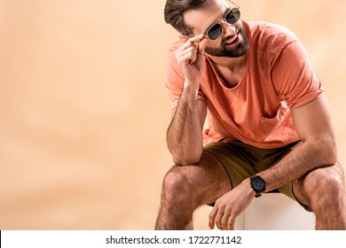 Happy Handsome Man In Shorts, Summer T-shirt And Sunglasses Sitting On White Cube On Beige