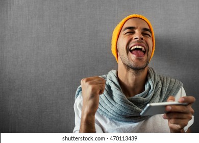 Happy Handsome Man With Telephone. Win In A Game. Wearing Orange Hat And Gray Scarf On Gray Background.