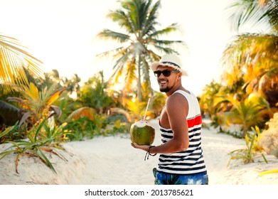 Happy handsome man wearing sunglasses and straw hat holding coconut on the tropical beach on sunny summer day during holidays vacation