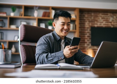 Happy Handsome Mature Asian Businessman Typing On Smartphone At Workplace At Laptop In Home Office Interior. Successful Business And Modern Technology, New Normal And Developing Marketing Strategy