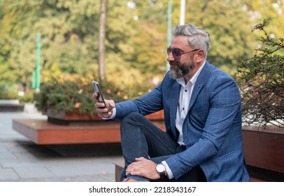 A Happy Handsome Mature Business Man Using Mobile Phone App Texting While Sitting On The Park Bench.