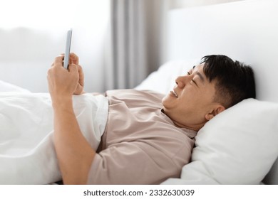 Happy handsome middle aged asian man wearing pajamas lying in bed at home, using cell phone and smiling, chatting with lover girlfriend, reading positive news, closeup
