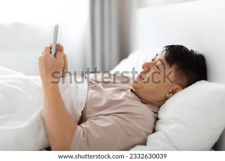 Happy handsome middle aged asian man wearing pajamas lying in bed at home, using cell phone and smiling, chatting with lover girlfriend, reading positive news, closeup