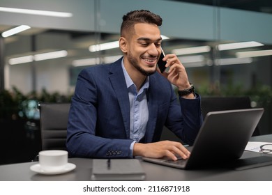 Happy handsome middle eastern businessman having phone conversation with client, sitting at worktable at office, typing on laptop keyboard, drinking coffee, copy space. Modern business concept