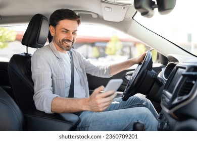 Happy handsome millennial guy driver using smartphone while driving car, using navigation system gps. Businessman chatting with friends or clients on mobile phone while going to office by auto - Powered by Shutterstock
