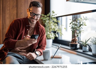 Happy handsome millennial man holding and using smartphone for typing, surfing the internet or writing text messages, sitting at table near window in the morning with cup of coffee and digital tablet. - Powered by Shutterstock