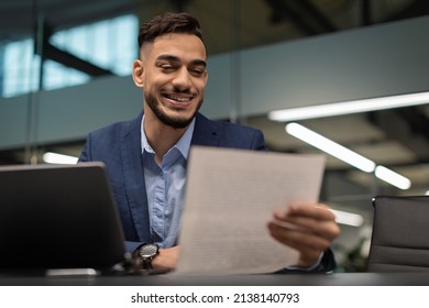 Happy Handsome Millennial Middle Eastern Businessman In Suit Sitting At Worktable In Front Of Laptop, Reading Marketing Report And Smiling, Modern Office Interior, Copy Space