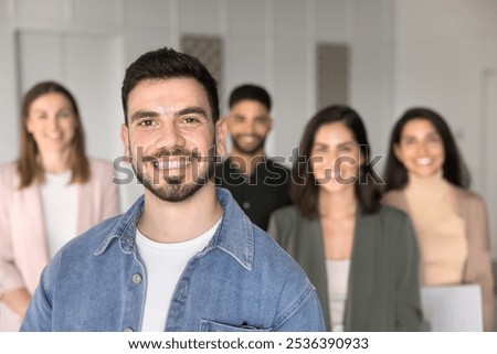 Happy handsome Muddle Eastern project leader man posing for business portrait, standing in front of multiethnic colleagues in background, smiling, laughing. Business company owner and staff shot