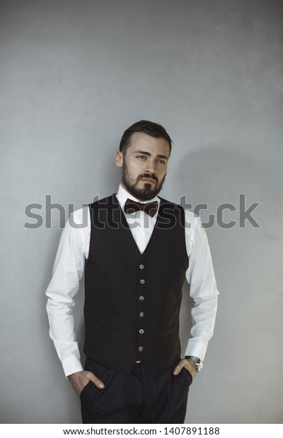 Happy handsome smiling\
man in a black vest and bow tie looking away. Portrait of a bearded\
attractive gentleman in elegant stylish clothes on gray grungy\
background.