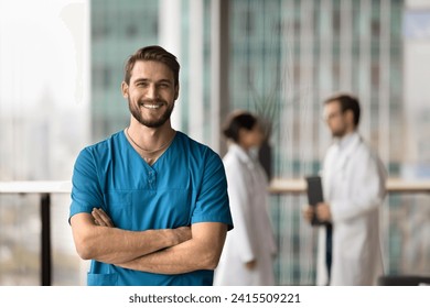 Happy handsome surgeon doctor man in blue uniform looking at camera with toothy smile, posing with hands crossed for professional portrait, standing in hospital hall space, laughing - Powered by Shutterstock