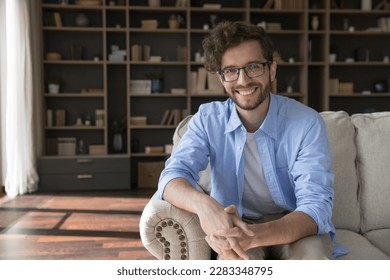 Happy handsome young 20s business man in glasses sitting on couch at home with bookshelves in background, looking at camera, smiling. Happy blogger, freelancer in eyeglasses head shot portrait - Powered by Shutterstock