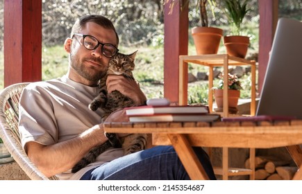 Happy handsome young adult businessman and his cat, man hugs his cute cat while working on laptop sitting at the work desk.