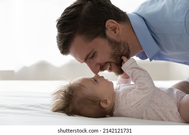 Happy handsome young dad calming, comforting baby resting on back in bed, kissing kid forehead with nose touches, expressing love, tenderness, care, caressing sleepy child. Fatherhood concept