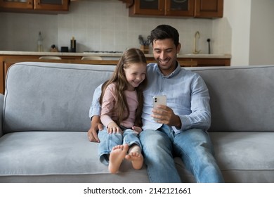 Happy Handsome Young Dad And Cute Preschool Daughter Kid Using Online Learning App On Mobile Phone, Making Funny Video Conference Call, Watching Content, Shopping On Internet, Sitting On Sofa