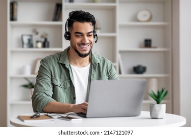 Happy Handsome Young Islamic Man With Beard In Headphones Looks At Laptop In Living Room Interior. Consultation Remotely, Social Distance, New Normal And Customer Support During Covid-19 Quarantine