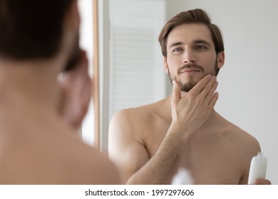 Happy handsome young man applying lotion or balm on stubble after-shaving or trimming, looking at mirror, touching beard. Guy enjoying male beauty care bathroom activity, holding cosmetic flask