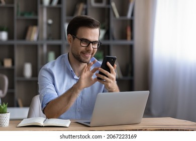 Happy handsome young man in eyewear looking at cellphone screen, reading message, or email, communicating distantly with client or playing games distracted from computer work in modern office.