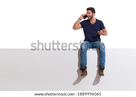 Happy handsome young man is sitting on a top, talking on the phone and looking away. Full length studio shot isolated on white.