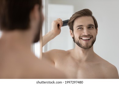 Feliz y apuesto hombre joven sin camisa peinando el cabello liso y liso, mirando en espejo, disfrutando de la actividad de cuidado de belleza, satisfecho con productos cosméticos de peluquería, corte de pelo, resultado de trabajo de barbero