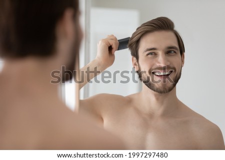 Happy handsome young shirtless man combing smooth straight hair, looking in mirror, enjoying beauty care activity, satisfied with haircare cosmetic products, haircut, barber work result