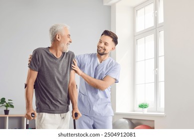 Happy health care worker with elderly man smiling and hugging in rehabilitation clinic. Young cheerful nurse helping senior patient to walk with his crutches. Physiotherapist helping old man in rehab - Powered by Shutterstock