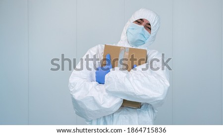 A happy health worker on protective suit hug a box of medical supplies with his arms in the hospital during pandemic caused by covid 19, coronavirus. The male nurse wears a surgical mask.