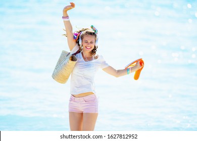 Happy Healthy 40 Year Old Woman In White T-shirt And Pink Shorts With Beach Straw Bag Listening To The Music With Headphones And Dancing On The Beach.