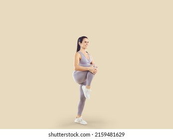 Happy Healthy Beautiful Woman In Comfortable Sports Clothes Having Fitness Workout. Fit Young Lady In Lilac Crop Top And Yoga Pants Standing Isolated On Beige Background And Doing High Knee Exercise