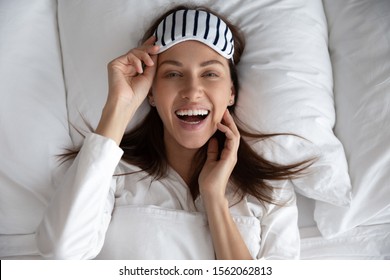 Happy Healthy Beautiful Young Woman Wear Sleeping Mask Awake After Sleep Laughing Lying In Comfortable Bed On Pillow, Smiling Lady Waking Up Enjoy Good Morning Looking At Camera, Portrait Top View