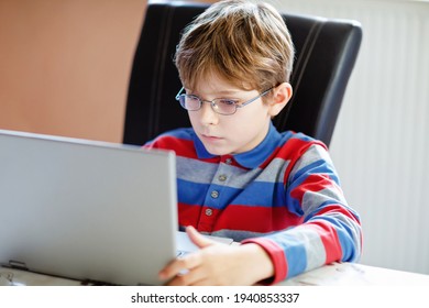 Happy Healthy Kid Boy With Glasses Making School Homework At Home With Notebook. Interested Child Writing Essay With Helping Of Internet. Concetrated Schoolchildren Concept