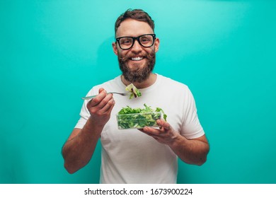 Happy healthy man eating salad wearing white t-shirt isolated over blue background. Healthy lifestyle concept - Shutterstock ID 1673900224