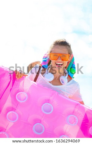 happy healthy middle age woman in white t-shirt and pink shorts with beach straw bag with inflatable mattress, headphones and orange sunglasses on the seacoast.