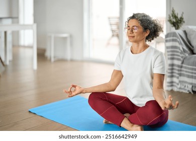 Happy healthy middle aged woman meditating at home. Serene mindful mature older lady in her 50s doing breathing exercises practicing meditation with eyes closed in modern house living room. Copy space - Powered by Shutterstock