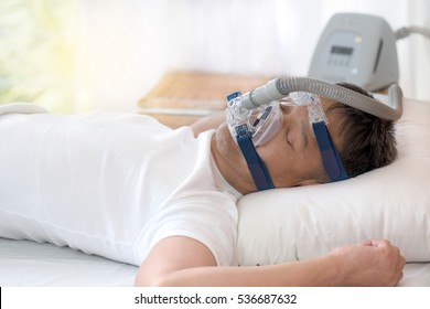 Happy and healthy senior man wearing cpap mask sleeping smoothly without snoring on his back open arms with blurred CPAP machine in background.Obstructive sleep apnea therapy ,close up  side view.
