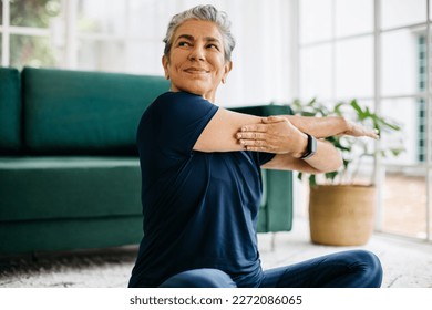 Happy and healthy senior woman doing a shoulder mobility exercise at home, working out with a cross arm stretch. Mature woman keeping herself active with yoga, maintaining her physical wellbeing. - Powered by Shutterstock