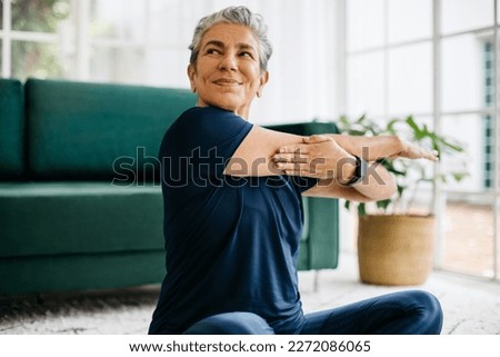 Happy and healthy senior woman doing a shoulder mobility exercise at home, working out with a cross arm stretch. Mature woman keeping herself active with yoga, maintaining her physical wellbeing.