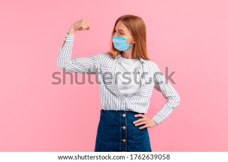 Happy healthy strong young woman in a medical protective mask on her face, showing biceps on her arm, on an pink background