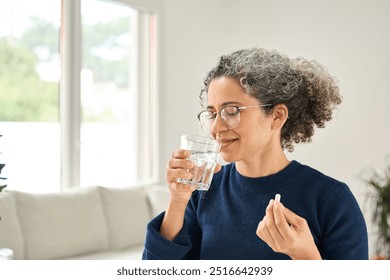 Happy healthy woman of middle age talking pill holding glass of water standing at home. Mature 50 years old lady taking vitamin health care supplement morning daily nutrition treatment concept. - Powered by Shutterstock