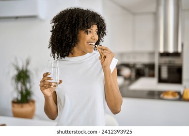 Happy healthy young adult African American woman model talking pill holding glass of water standing in the kitchen at home. Morning supplements vitamins nutrition treatment concept.