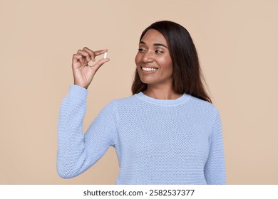 Happy healthy young adult African American woman holding vitamin in hand looking at pill standing isolated on beige background advertising beauty supplements nutrition treatment concept.