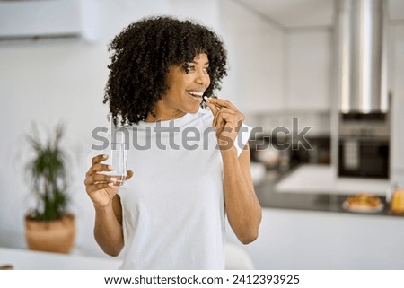 Happy healthy young adult African American woman model talking pill holding glass of water standing in the kitchen at home. Morning supplements vitamins nutrition treatment concept.