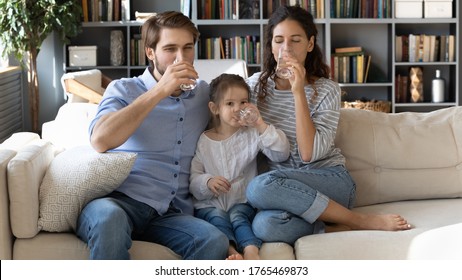 Happy healthy young family drinking pure mineral water together, thirsty dehydrated mother and father with cute little daughter holding glasses, sitting on couch in living room, healthy lifestyle - Shutterstock ID 1765469873