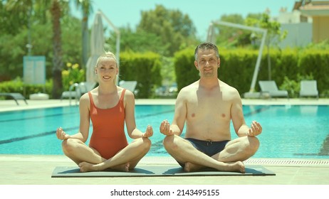 Happy Healthy Young Husband And Wife Getting Ready To Practice Yoga Together By The Pool, Sitting In Lotus Pose And Looking At Each Other Lovingly. Portrait Of A Happy Couple Relaxing On Vacation. 