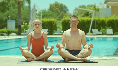 Happy Healthy Young Husband And Wife Getting Ready To Practice Yoga Together By The Pool, Sitting In Lotus Pose And Looking At Each Other Lovingly. Portrait Of A Happy Couple Relaxing On Vacation. 